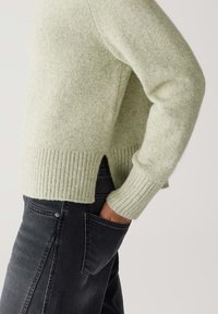 Person wearing a light green ribbed sweater with side slits and black jeans, hand tucked into the back pocket, on a neutral background.