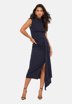 Žena s dlhými tmavými vlasmi nosí čierne midi šaty bez rukávov so štýlom na boku a záhybným detailom, v kombinácii s čiernymi lodičkami na členkový remienok.