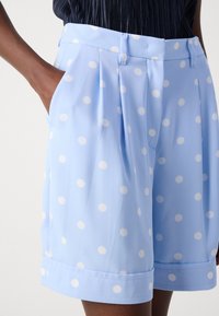 Light blue pleated shorts featuring white polka dots, a structured waistband, and side pockets. Soft, smooth fabric with a relaxed fit.