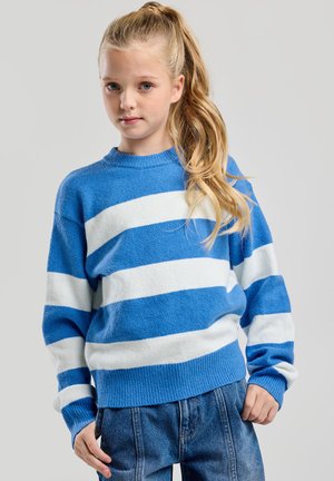 Jong meisje met een lange blonde paardenstaart, gekleed in een blauw-wit gestreepte trui en een blauwe spijkerbroek, staand tegen een effen achtergrond.