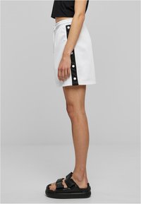 Pantalones cortos blancos con panel lateral negro, botones de presión y cintura elástica. Textura suave, diseño hasta la rodilla. Sandalias negras completan el conjunto.
