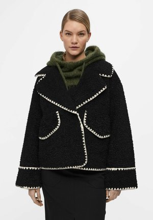 Schwarzer strukturierter Mantel mit breitem Revers, hellem Paspel und zwei Fronttaschen. Getragen über einem grünen Strickhoodie mit weichem Aussehen.