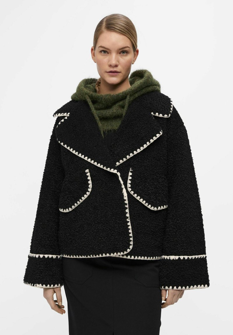 Cappotto nero con texture, colletto ampio, dettagli in crema, e due tasche frontali. Indossato sopra un hoodie verde lavorato a maglia, caratterizzato da un aspetto morbido.