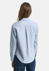 Donna con capelli lunghi che indossa una camicia blu chiaro con bottoni e jeans blu scuro, vista di spalle su uno sfondo semplice.