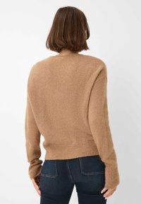 Beige, zacht gebreide trui met verlaagde schouders, geribbelde zoom en manchetten, en een ronde halslijn. Draagt met donkere jeans tegen een effen achtergrond.