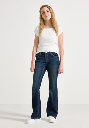Vitt kortärmat tröja i kombination med mörkblå utsvängda jeans. Enkel design med vita sneakers, stående mot en neutral bakgrund.