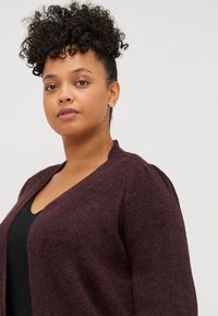 Cardigan bordeaux con scollo a V, in tessuto morbido e texture delicate, con pieghe sottili sulle spalle. Indossato sopra un top nero, mette in risalto la sua vestibilità comoda.