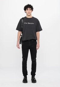 Camiseta gráfica negra de manga corta, que presenta el texto "The Rougher" en un contraste ligero. Combinada con jeans negros desgastados y zapatos negros.