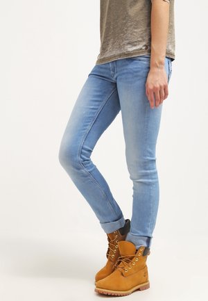 Ljusblå skinny jeans med en blekt finish, tillsammans med bruna snörkängor. Överdelen är en strukturerad olivgrön t-shirt med kort ärm.