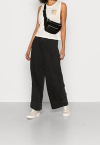 Pantalones anchos negros, camiseta sin mangas blanca con gráfico y bolso bandolera negro. Zapatillas de color claro con cordones. Textura suave y estilo casual.