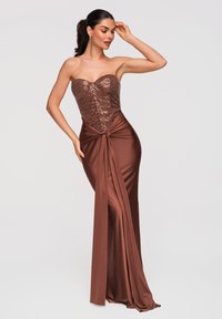 Femme posant dans une robe bustier cuivre avec corsage à paillettes et détail de ceinture nouée, portant de longues boucles d'oreilles, sur fond gris uni.