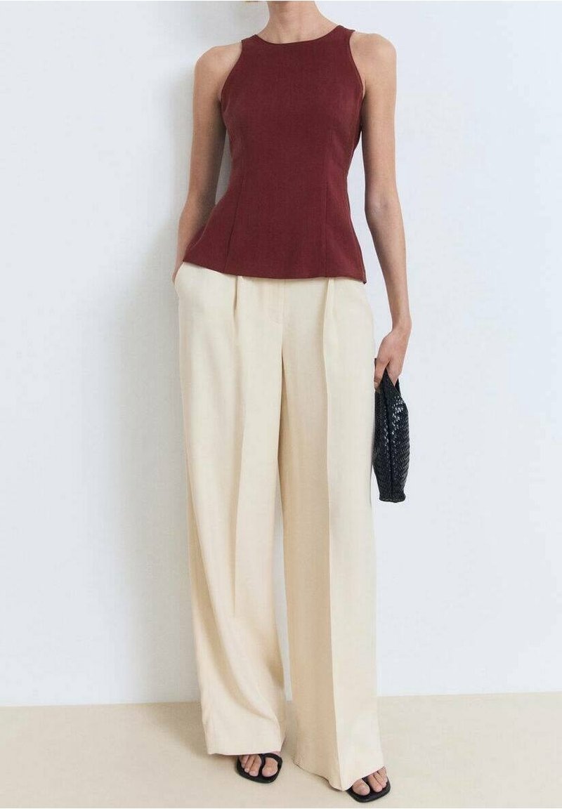 Femme portant un haut bordeaux sans manches, un pantalon large crème, des sandales noires, tenant une petite pochette noire texturée contre un mur blanc.