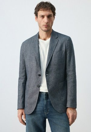 Uomo che indossa un blazer grigio sopra una camicia bianca abbinata a jeans blu, in piedi davanti a uno sfondo chiaro uniforme.
