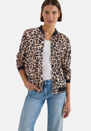 Jeune femme portant une veste zippée beige et noire à imprimé léopard sur une chemise blanche et un jean bleu clair, avec une main dans la poche.
