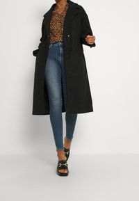 Cappotto trench nero con colletto a rever, design doppiopetto, indossato sopra un top in stampa leopardata e jeans skinny blu, abbinato a sandali neri.