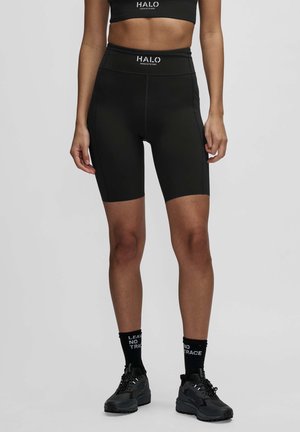 Zwarte high-waisted fietsshorts gemaakt van rekbare stof, met een logo tailleband en een gestroomlijnd ontwerp, gecombineerd met zwarte sneakers en sokken.