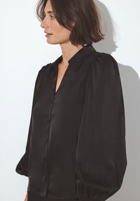 Blusa nera in raso con colletto classico, chiusura a bottoni frontale e maniche lunghe a sbuffo, dal tessuto morbido e dal fit rilassato.