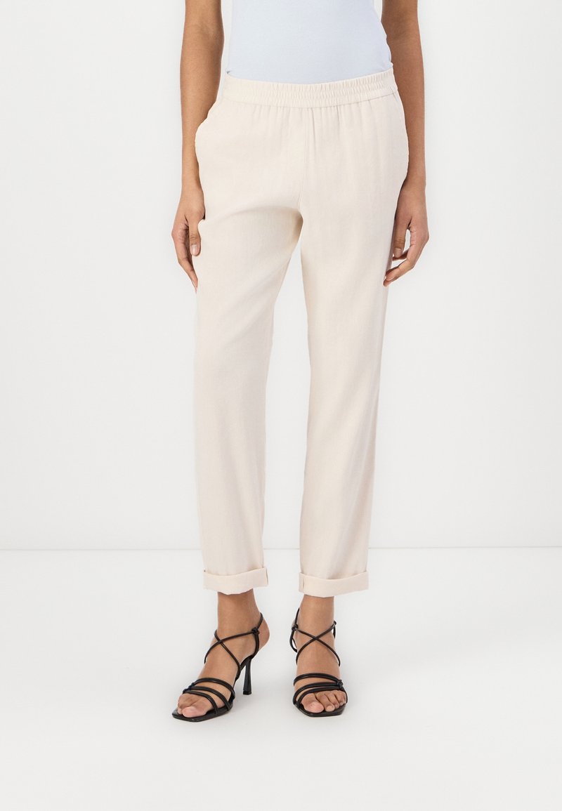 Pantaloni beige chiaro con vita elastica, gambe affusolate e risvolti. Indossati con sandali neri con tacchi alti e cinturini. Tessuto morbido.