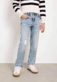 Jeans in denim azzurro chiaro, dritti e con dettagli strappati. Abbinati a sneaker bianche con accenti beige e un maglione corto a righe blu navy.