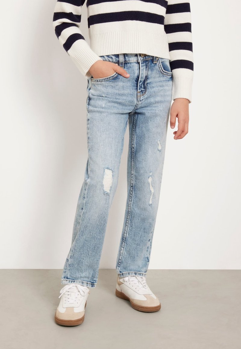 Jeans in denim azzurro chiaro, dritti e con dettagli strappati. Abbinati a sneaker bianche con accenti beige e un maglione corto a righe blu navy.