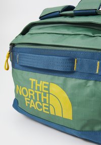 Green and blue duffel bag with yellow logo. Features a textured fabric, sturdy zippers, and reinforced handles for durability.