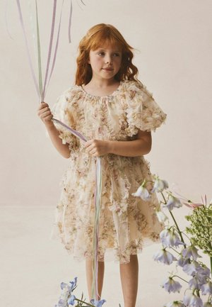 Junges Mädchen mit roten Haaren, das ein gerafftes, geblümtes Kleid trägt und Pastellfarbene Luftballons hält, steht in der Nähe von blau-weißen Blumen.