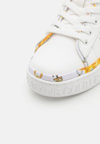 Witte sneaker met een gestructureerde zool, stoffen accenten met kleurrijke patronen en platte veters. "JEANS COUTURE" is in de zijkant geperst.