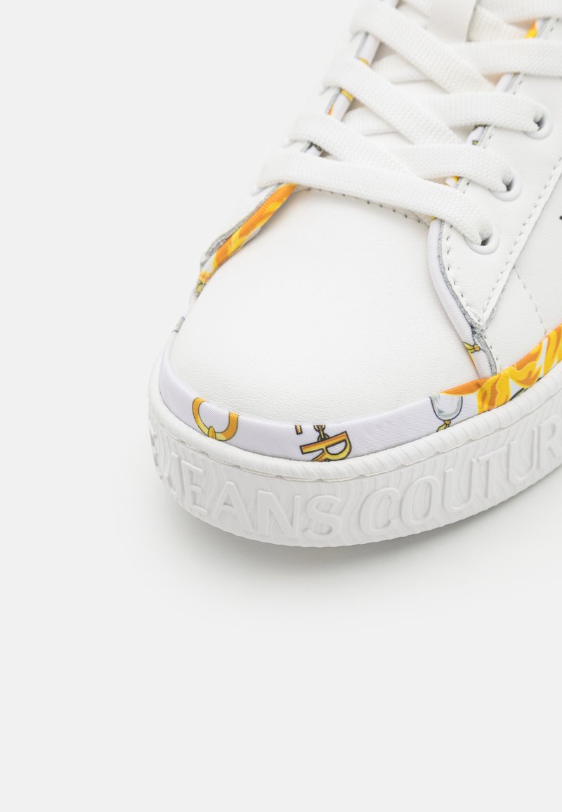 Zapatilla blanca con suela texturizada, acentos de tela con patrones coloridos y cordones planos. "JEANS COUTURE" grabado en el costado.