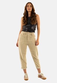 Beige ribfluwelen broek met hoge taille, voorzakken en opgerolde manchetten, gecombineerd met een zwart mouwloos topje en lage sneakers.