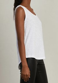 Bílý bezrukávový top s kulatým výstřihem, vyrobený z lehké strukturované tkaniny. Boční lem je mírně delší vzadu.