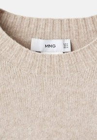 Guler de pulover din tricot bej cu striații, având o etichetă albă care arată marca "MNG" și mărimea medie pentru piețele din EUR, SUA și MEX.