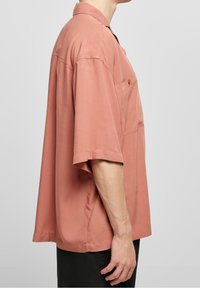Camicia a maniche corte, con colletto, in rosa polveroso, realizzata in tessuto liscio. Presenta una tasca sul petto e una vestibilità morbida e oversize.