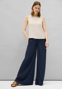 Top sin mangas beige con un acabado texturizado, combinado con pantalones de pierna ancha en color azul marino. La modelo lleva sandalias marrones con recortes decorativos.