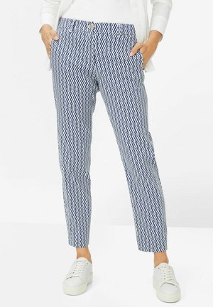 Femme portant un pantalon slim à motif géométrique bleu et blanc, mains dans les poches et baskets blanches, debout sur un fond blanc.