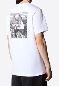 Person wearing a white t-shirt with a black and white mountain and forest graphic and "The North Face" logo on the back.