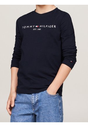 Person iført en marineblå langærmet Tommy Hilfiger-t-shirt med hvid logo-tekst og blå denim-jeans, hånd i lommen, mod en ensfarvet baggrund.