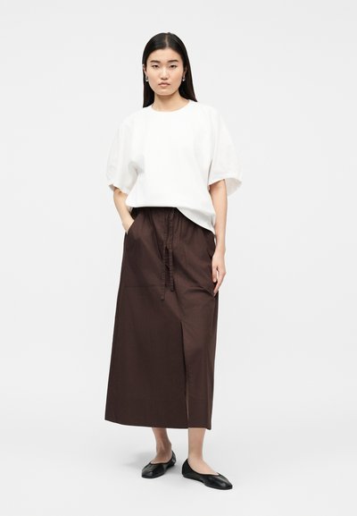 Γυναίκα όρθια που φορεί λευκό μπλουζάκι με κοντά μανίκια, σκουρόχρωμη καφέ μίντι φούστα με ψηλή μέση και σχίσιμο μπροστά, μαύρα μπαλαρίνες και ασημένια σκουλαρίκια στεφάνια.