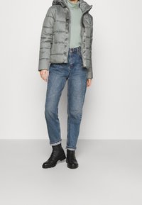 Šedá péřová bunda s kapucí, světlá zelená rolák, modré džínové kalhoty a černé šněrovací boty, s volným střihem a strukturovanou tkaninou.