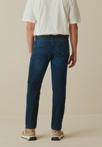 Vaqueros de mezclilla azul oscuro con un corte recto, que cuentan con dos bolsillos traseros y detalles de costura sutiles, combinados con zapatos beige y una camiseta blanca.