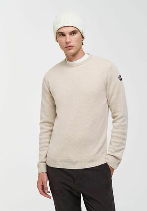 Junger Mann mit beigem Pullover und weißer Strickmütze, steht mit einer Hand in der Tasche vor einem schlichten hellen Hintergrund.