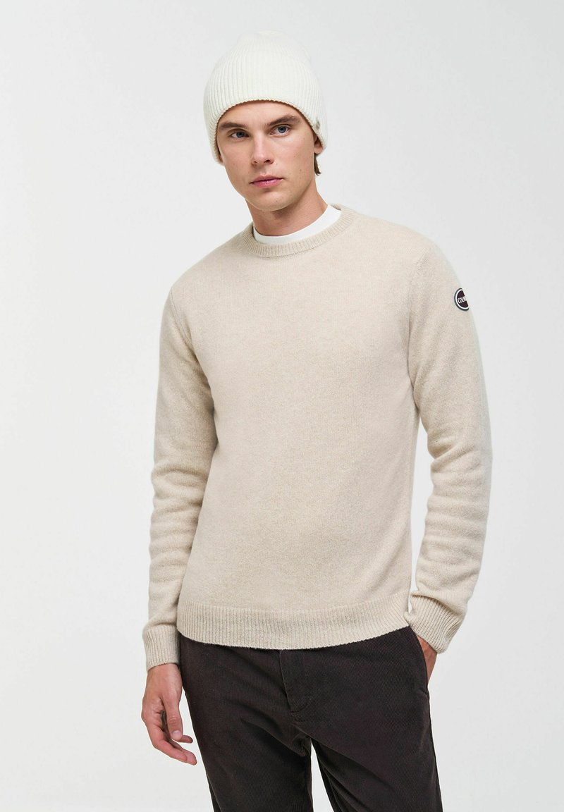 Jeune homme portant un pull beige et un bonnet en maille blanc, debout avec une main dans la poche sur un fond clair uni.