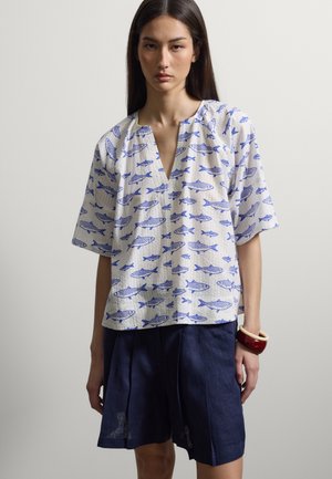 Femme portant un chemisier blanc ample avec un imprimé de poissons bleus et un short plissé bleu marine, debout devant un fond clair uni.