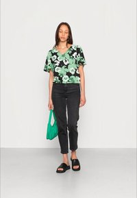 Camisola de manga curta floral em preto com padrões verdes e brancos, combinada com jeans pretos e sandálias deslizantes pretas. Segura uma bolsa verde.