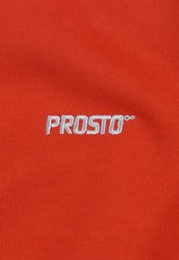 Czerwona tkanina o gładkiej fakturze z wyhaftowanym logo "PROSTO" w białym kolorze. Wzór jest prosty i umieszczony w centrum tkaniny.