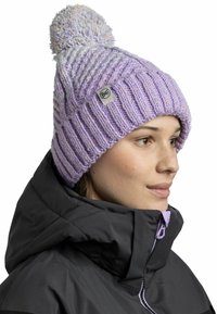 Purple knit beanie with a ribbed design, featuring a fluffy pom-pom on top and a small brand tag. Worn with a black jacket, showing collar details.