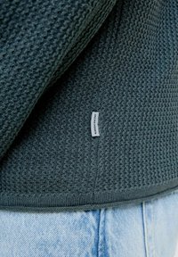 Pull en maille texturée vert foncé avec un motif tissé, doté d'un ourlet côtelé et d'une petite étiquette sur le côté. Porté sur un jean en denim bleu clair.