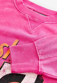 Sweat-shirt à col rond rose avec col et poignets côtelés, présentant un texte imprimé en jaune et noir usé sur le devant.