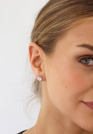 Ohrringe aus Roségold mit runden Kristallmitte, in einer strukturierten Goldbasis eingesetzt, Nahaufnahme am Ohr eines Modells.