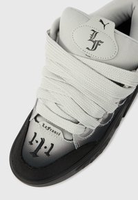 Κοντινό πλάνο σε γκρι και μαύρο αθλητικό παπούτσι με φαρδιές, υφασμένες κορδόνια, το κείμενο "LaFrance" και ένα ραμμένο οβάλ λογότυπο με τα γράμματα "LF" και το σύμβολο της Puma στη γλώσσα του παπουτσιού.