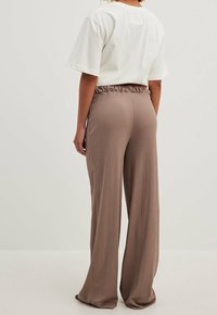 Weißes T-Shirt mit kurzen Ärmeln und einem quadratischen Aufnäher auf dem Rücken, kombiniert mit braunen, gerippten, weit ausgestellten Hosen mit elastischem Bund.